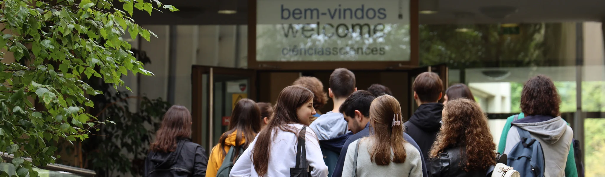 estudantes entram nas instalações da Faculdade de Ciências da Universidade do Porto; por cima da porta, uma placa tem escrito "bem-vindos. welcome"